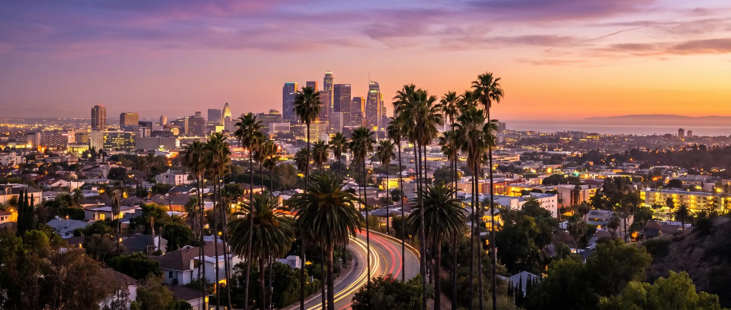 The Downtown Los Angeles skyline at dusk, seen across palm trees and hillside neighborhoods